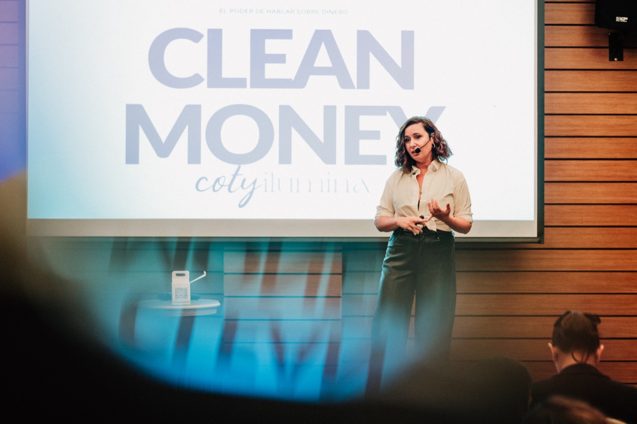 Coty de pie en el escenario, con micrófono, dando su charla Clean Money frente a la pantalla